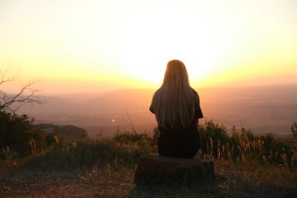 Person sitting on rock at crest of hill looking out at sunset.